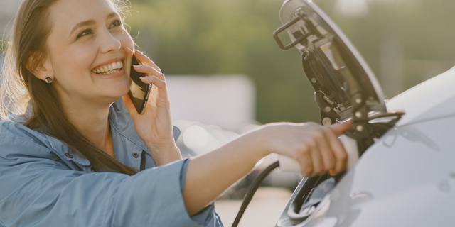 Mujer carga un vehículo eléctrico mientras conversa, feliz, por teléfono