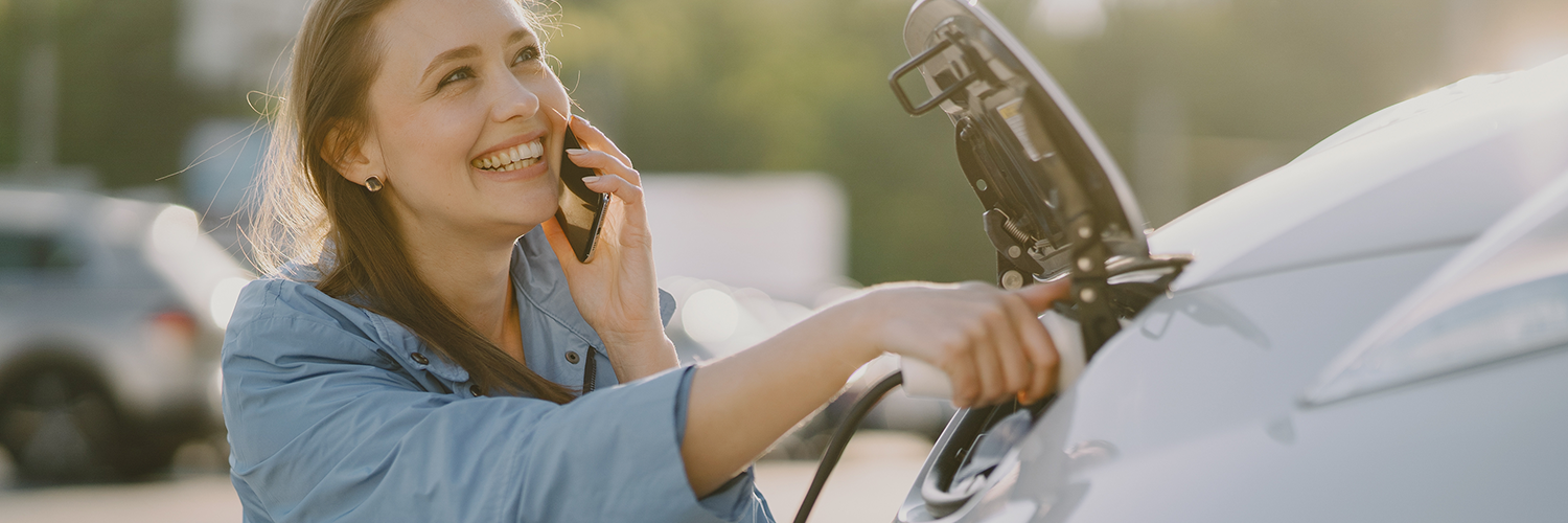 Mujer carga un vehículo eléctrico mientras conversa, feliz, por teléfono